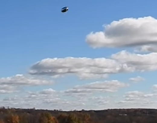 Photo d'un ballon en Mylar se tenant horizontalement dans le ciel, avec la partie supérieure reflétant le ciel et la partie inférieure sombre et légèrement rougeâtre, du fait qu'elle reflète le sol